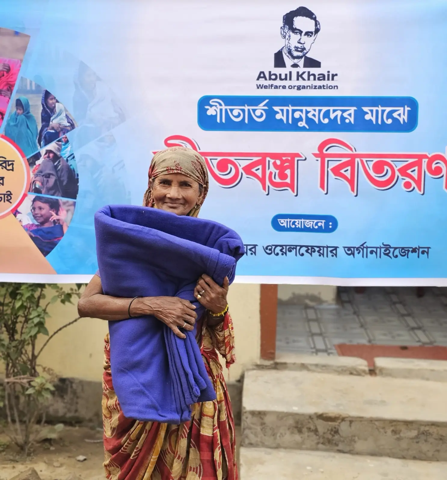 A woman holds the blanket she has received in front of the AKWO winter distribution banner, January 2025