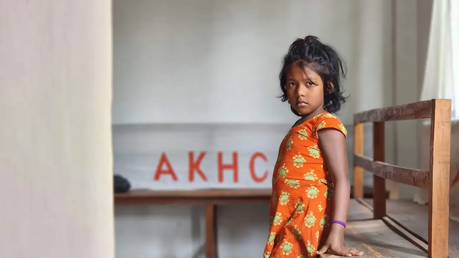 A young girl stands beside a bench under the AKHC sign at the Baishmouja clinic