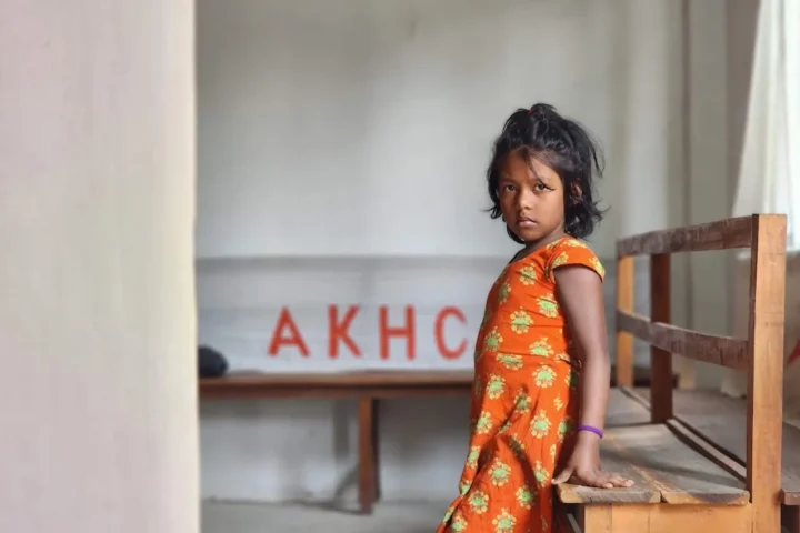 A young girl stands beside a bench under the AKHC sign at the Baishmouja clinic