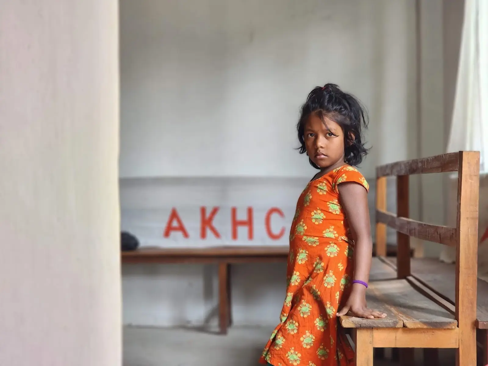A young girl stands beside a bench under the AKHC sign at the Baishmouja clinic