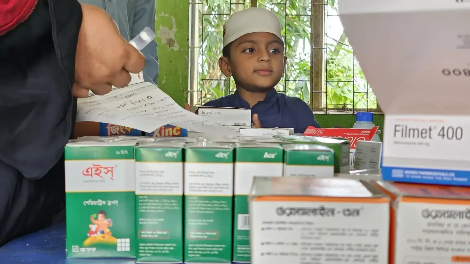 Boxes of free medicine stacked on the dispensing table at the Baishmouja clinic while a boy waits at the counter