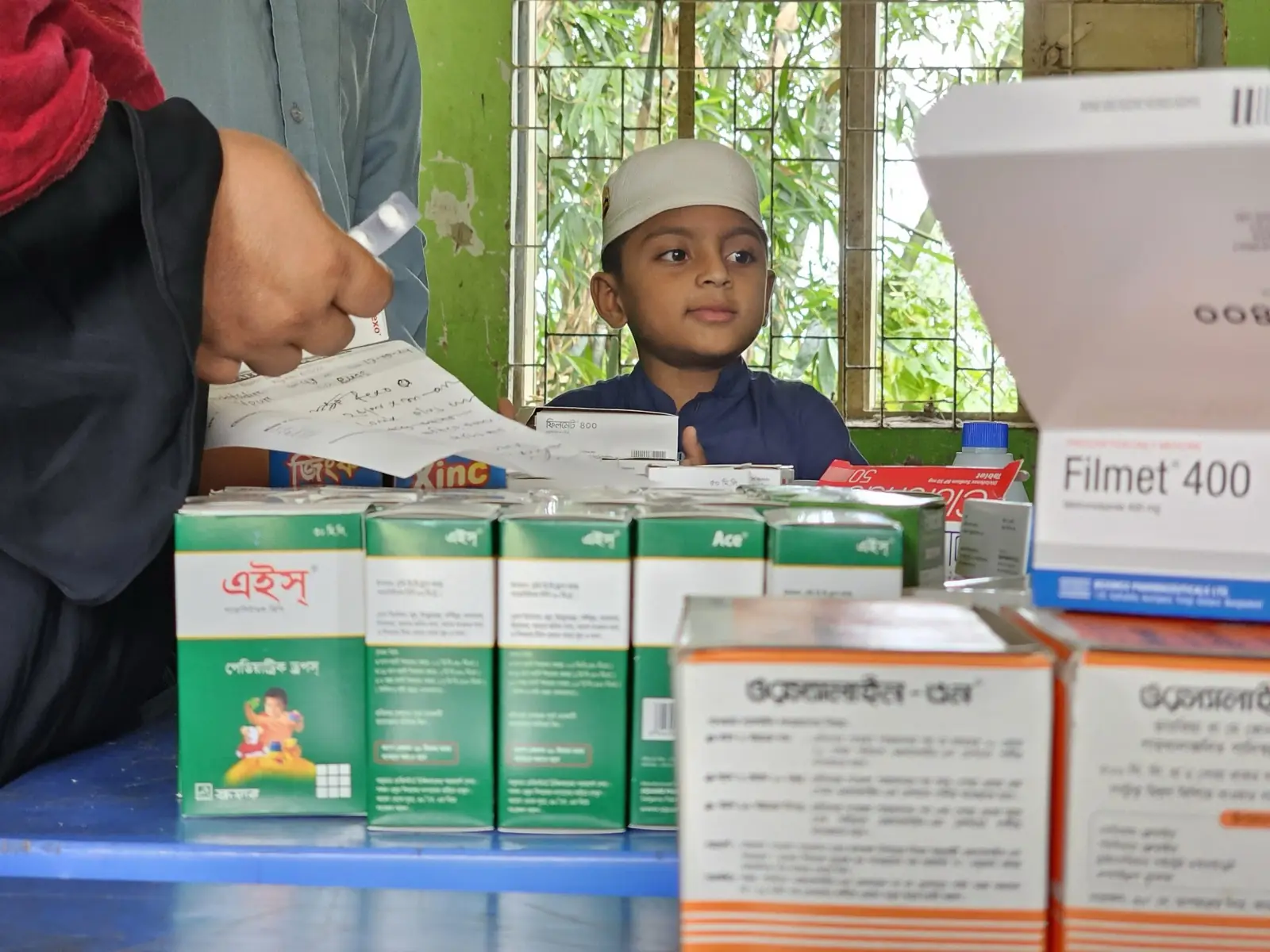 Boxes of free medicine stacked on the dispensing table at the Baishmouja clinic while a boy waits at the counter