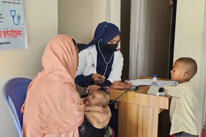 An AKWO health assistant records a woman's blood pressure while her child stands beside the desk