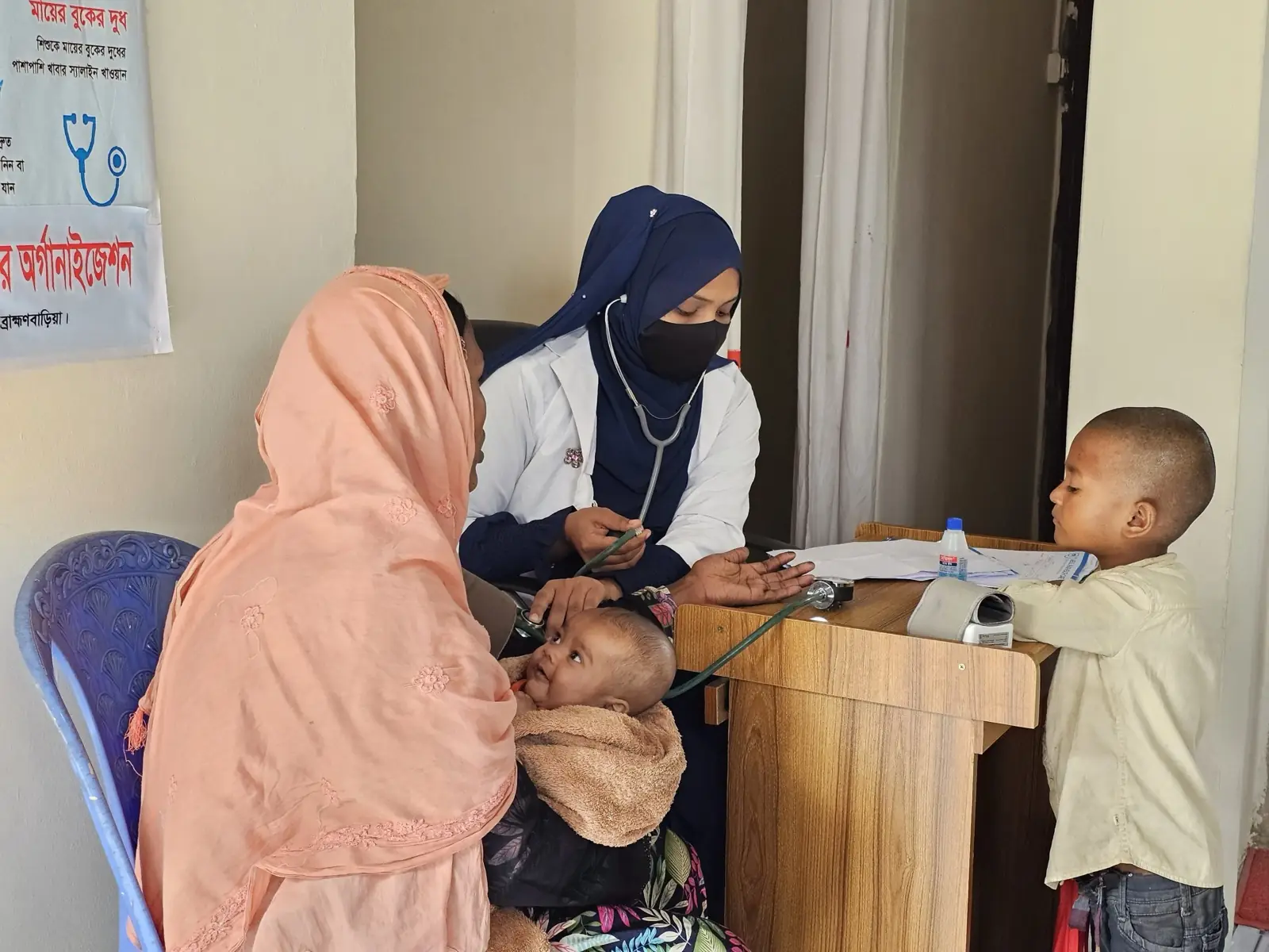 An AKWO health assistant records a woman's blood pressure while her child stands beside the desk