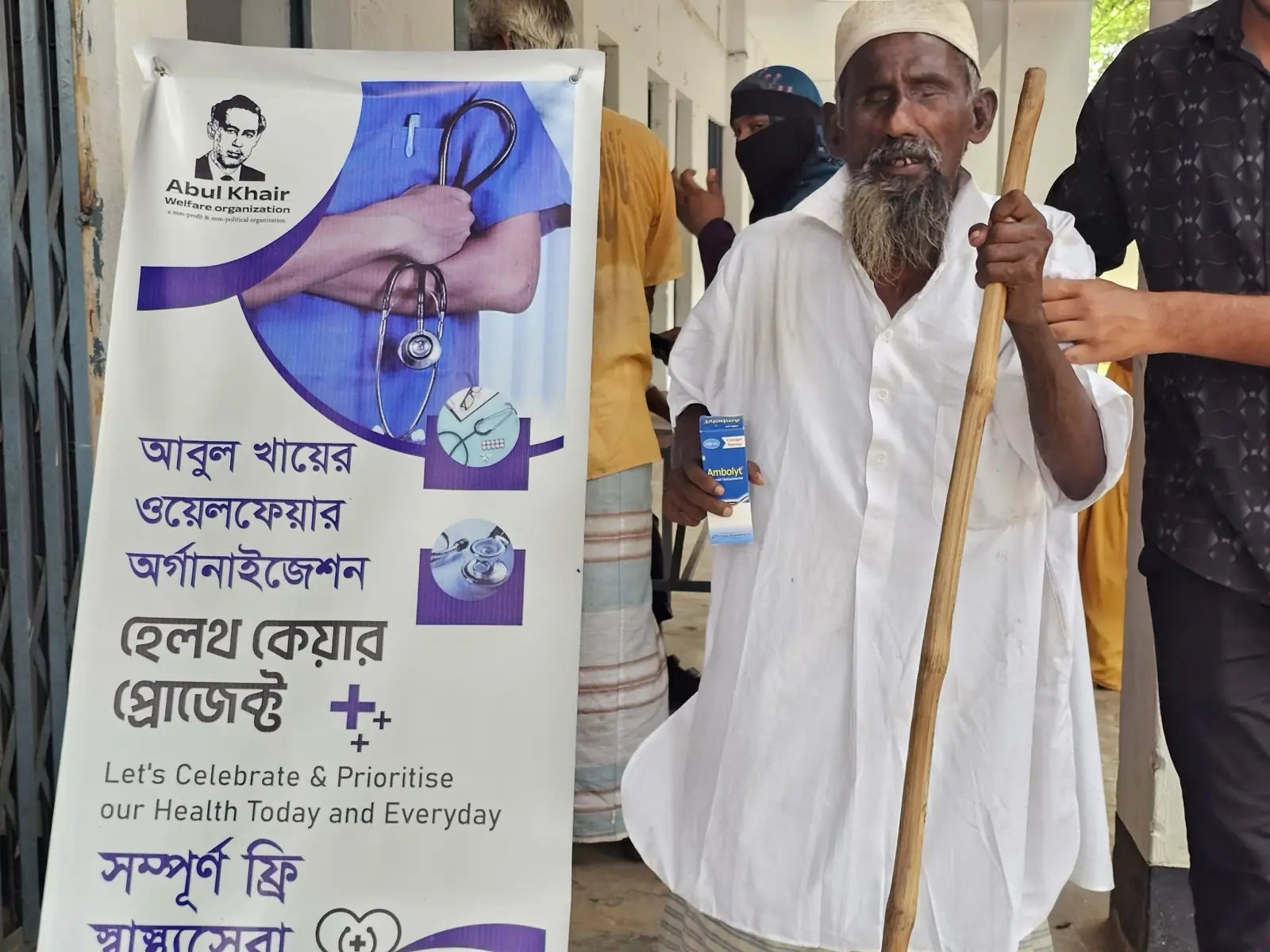 An elderly man with a walking stick arrives at the free clinic beside the AKWO health care banner